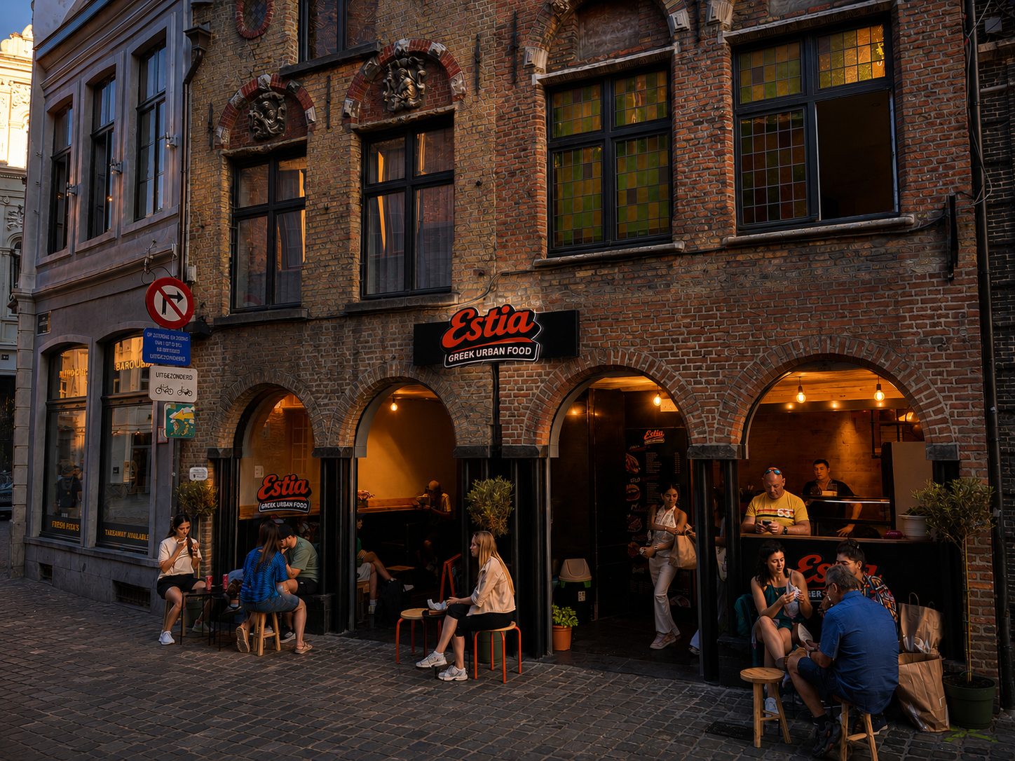 ESTIA exterior in Bruges at evening with the sign lit and guests seated outside